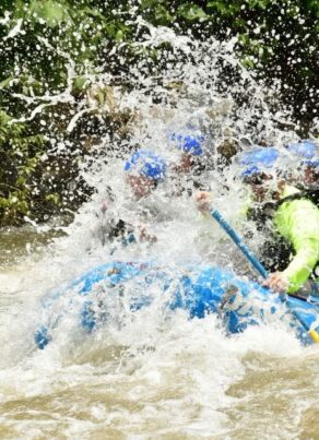 A group of people wearing helmets and life jackets paddle a blue raft through splashing whitewater rapids, surrounded by trees and rocks.