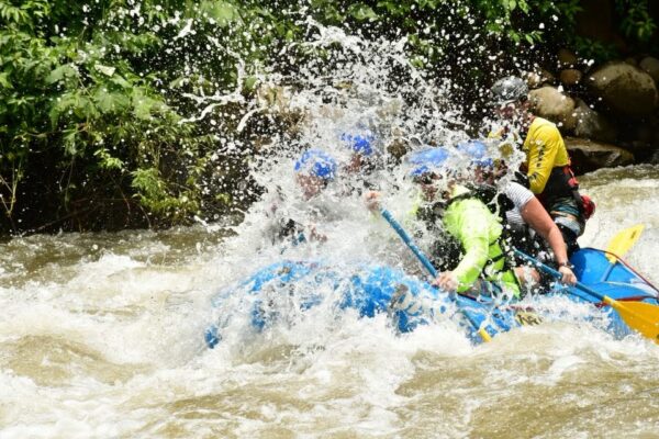 A group of people wearing helmets and life jackets paddle a blue raft through splashing whitewater rapids, surrounded by trees and rocks.