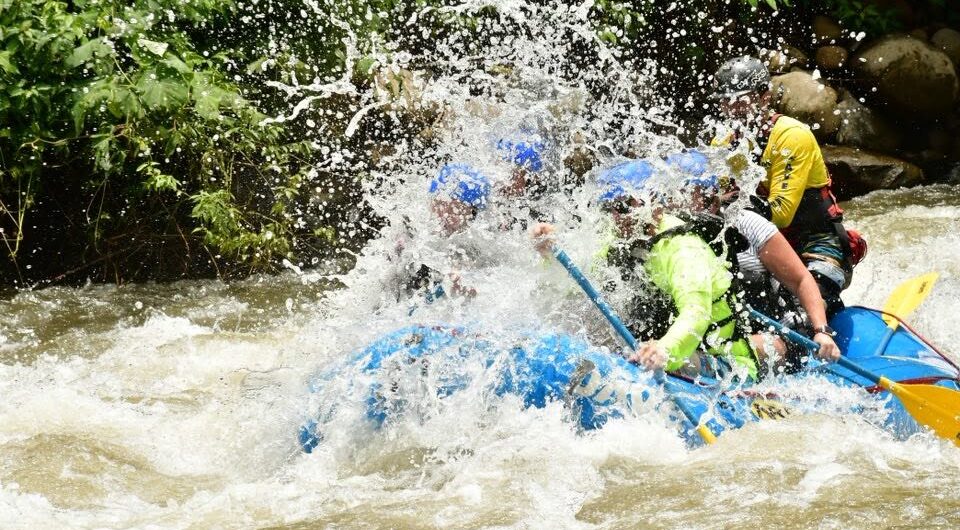 A group of people wearing helmets and life jackets paddle a blue raft through splashing whitewater rapids, surrounded by trees and rocks.