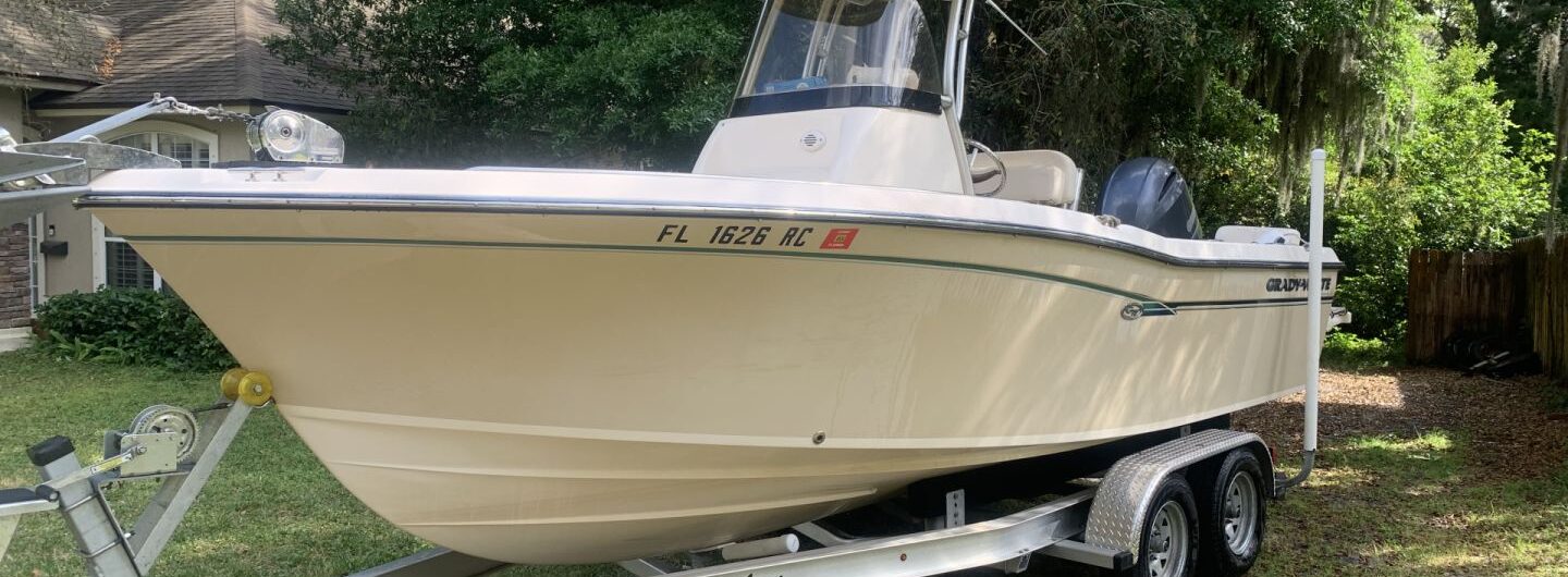 A beige center console fishing boat on an “Advanced Boat Trailers” trailer is parked on grass beside a house, surrounded by trees, with a Florida registration number on the hull.