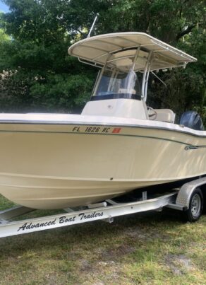 A beige center console fishing boat on an “Advanced Boat Trailers” trailer is parked on grass beside a house, surrounded by trees, with a Florida registration number on the hull.