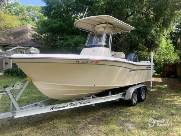 A beige center console fishing boat on an “Advanced Boat Trailers” trailer is parked on grass beside a house, surrounded by trees, with a Florida registration number on the hull.