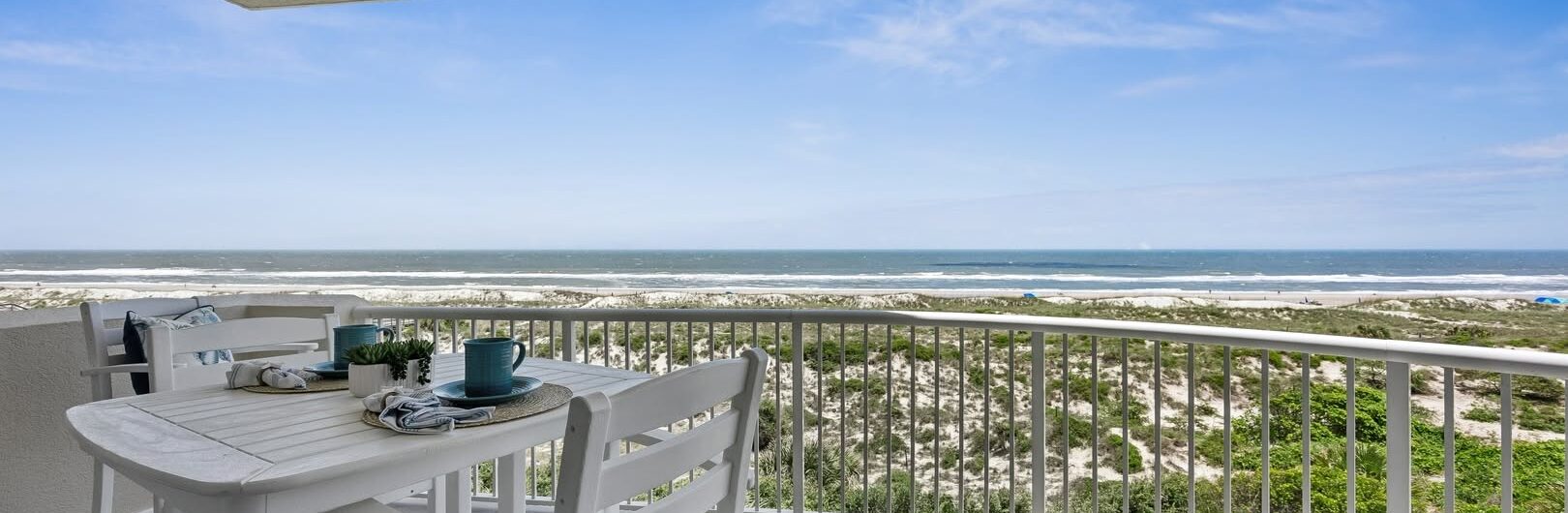 A white balcony with a table and chairs overlooks sand dunes and the ocean under a blue sky with scattered clouds. Pillows and mugs are on the table, creating a relaxing seaside atmosphere.