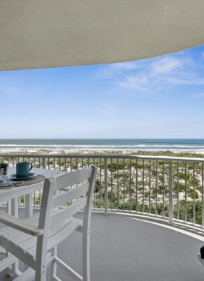 A white balcony with a table and chairs overlooks sand dunes and the ocean under a blue sky with scattered clouds. Pillows and mugs are on the table, creating a relaxing seaside atmosphere.