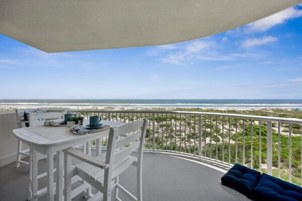 A white balcony with a table and chairs overlooks sand dunes and the ocean under a blue sky with scattered clouds. Pillows and mugs are on the table, creating a relaxing seaside atmosphere.
