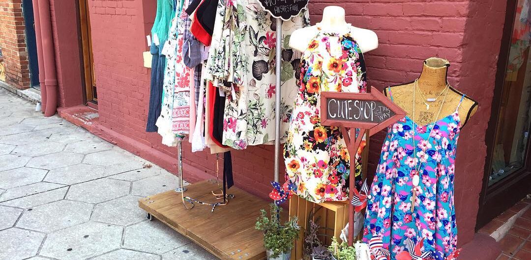 Outdoor clothing display in front of a red brick wall, featuring dresses on hangers, two mannequins in colorful floral dresses, potted plants, and a sign reading “CLUE SHOPPE.”.