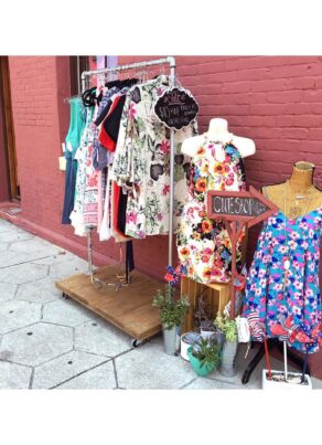 Outdoor clothing display in front of a red brick wall, featuring dresses on hangers, two mannequins in colorful floral dresses, potted plants, and a sign reading “CLUE SHOPPE.”.