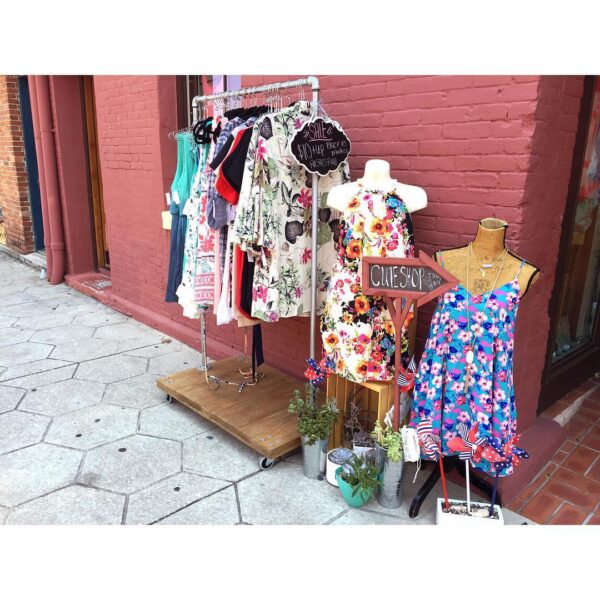 Outdoor clothing display in front of a red brick wall, featuring dresses on hangers, two mannequins in colorful floral dresses, potted plants, and a sign reading “CLUE SHOPPE.”.