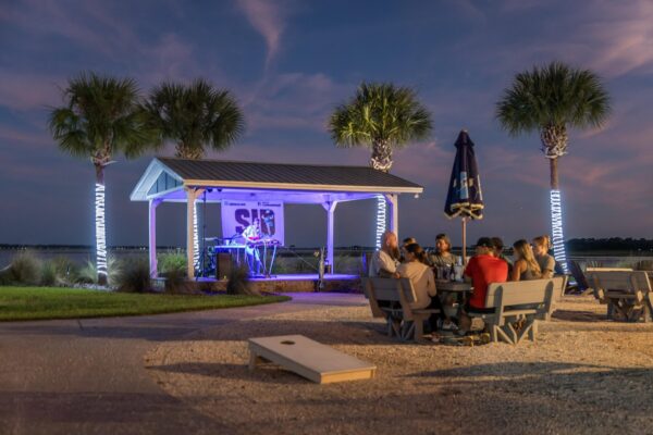 People sit on benches under palm trees, watching a musician perform on a lit outdoor stage at dusk. An umbrella and a cornhole game are nearby; the setting feels relaxed by the waterfront.