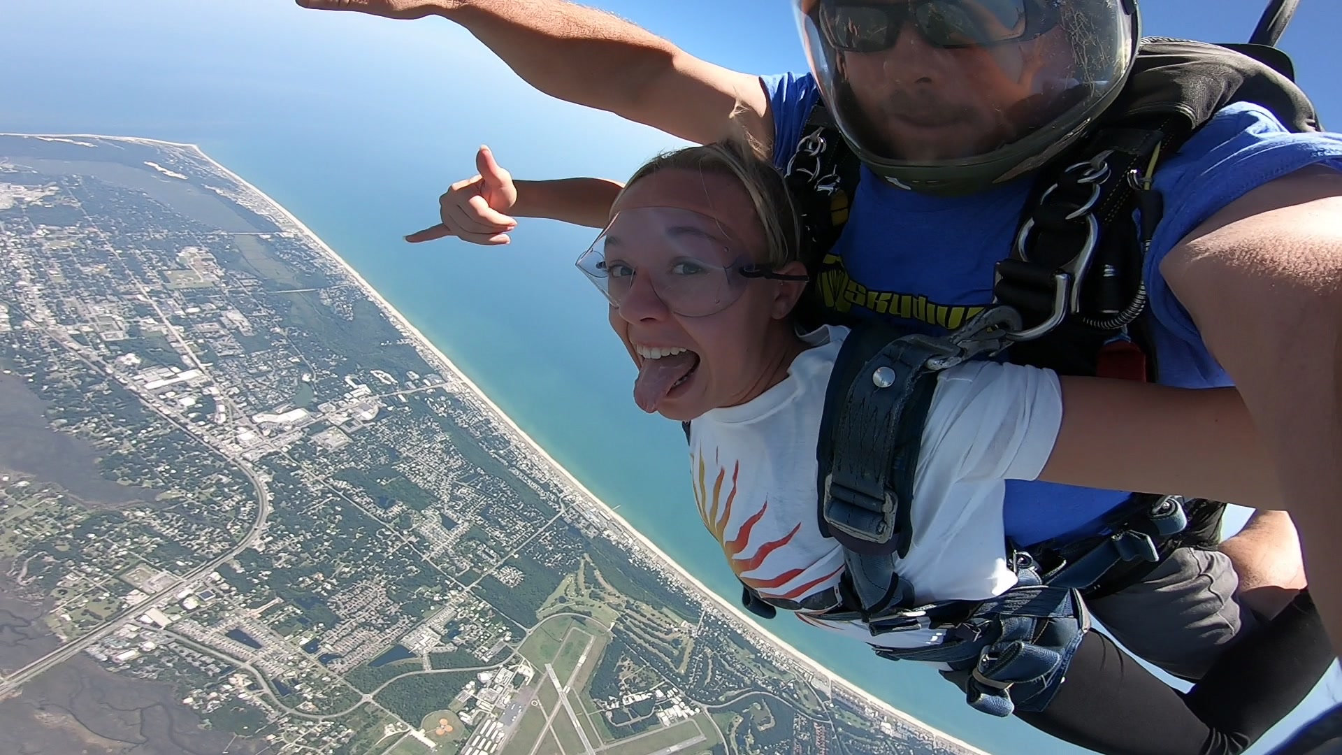 Two people are tandem skydiving high above a coastline and town. The person in front is smiling, sticking out their tongue, and giving a shaka hand sign, while the instructor holds onto them from behind.