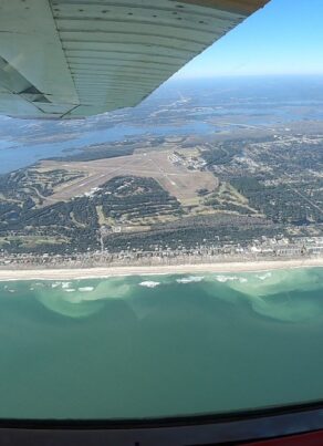 Aerial view from an airplane showing a coastline with sandy beach, light green and blue ocean waves, a town, an airport runway, and distant landmasses under a clear blue sky.