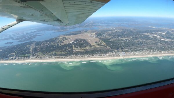 Aerial view from an airplane showing a coastline with sandy beach, light green and blue ocean waves, a town, an airport runway, and distant landmasses under a clear blue sky.