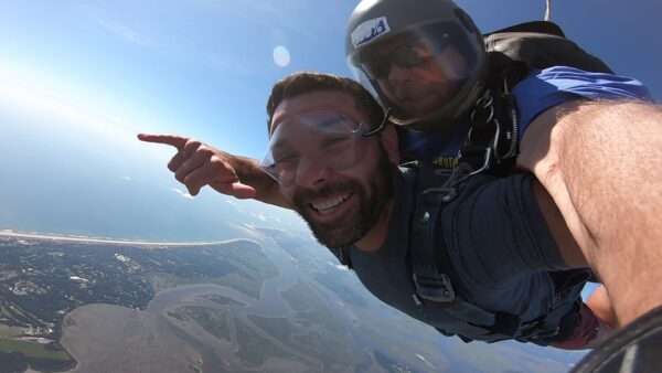 Two people tandem skydiving high above a coastal landscape. The person in front is smiling and pointing towards the horizon, while the instructor behind them holds on tightly. Both are wearing helmets and goggles.
