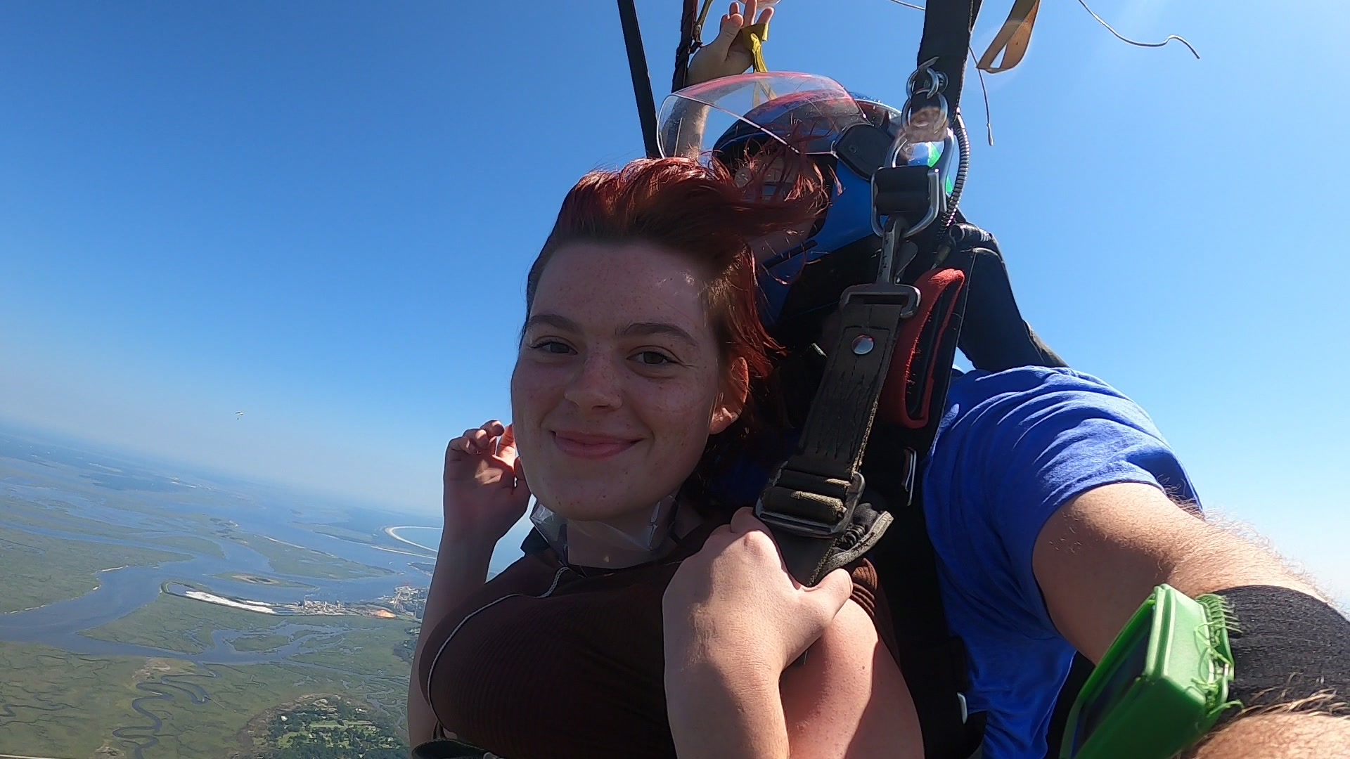 A woman with reddish hair smiles while tandem skydiving, strapped to an instructor. The blue sky and sprawling green landscape with rivers are visible far below them.