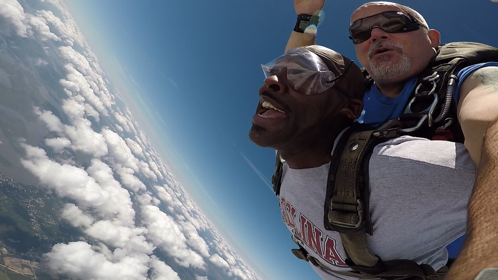 Two men tandem skydiving in mid-air, both wearing goggles and harnesses. The man in front has his mouth open in excitement; the instructor behind him has one arm raised. Blue sky and clouds fill the background.