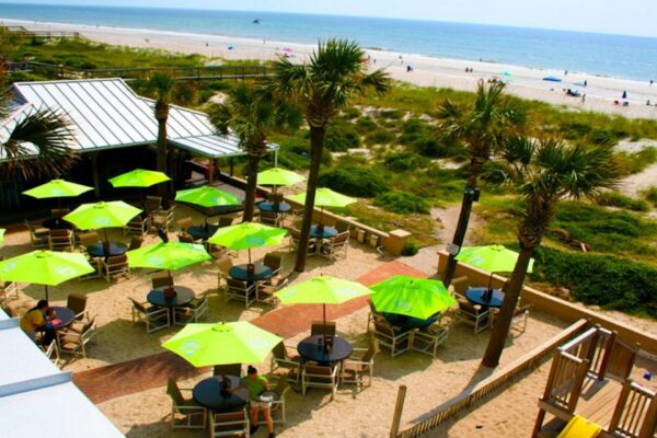 Aerial view of a beachside outdoor restaurant with green umbrellas, palm trees, and round tables on sandy ground. The beach and ocean are visible in the background with people and umbrellas along the shore.