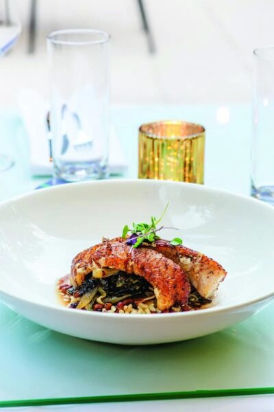 A white plate with a seared fish fillet garnished with microgreens, served over a bed of grains and vegetables. A gold candle holder and clear glasses are in the background on a glass table.