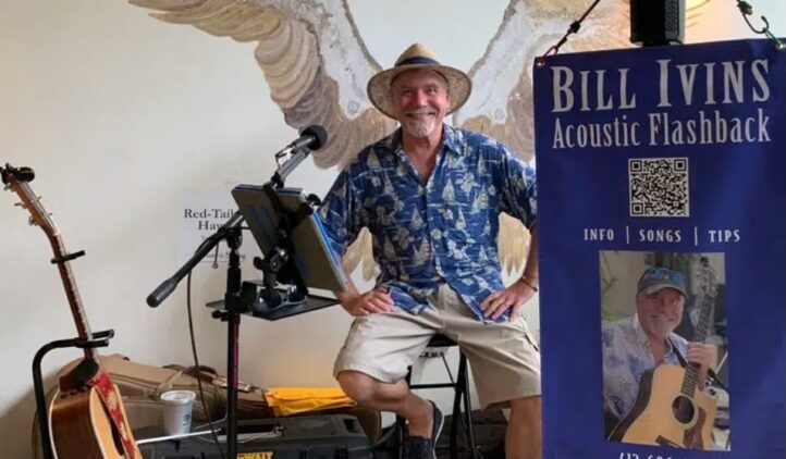 A smiling musician in a blue Hawaiian shirt and straw hat sits on a stool with instruments nearby. A sign reads Bill Ivins Acoustic Flashback with contact info and a photo of him holding a guitar. A mural of wings is on the wall behind him.