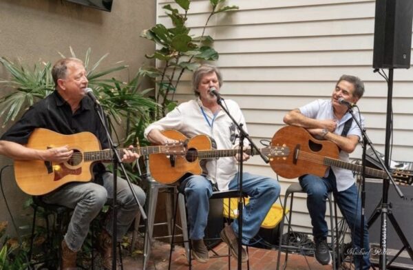 Three men sit and play acoustic guitars, singing into microphones on stands. They are performing together in an outdoor patio area lined with plants and white siding. All appear to be enjoying the music.