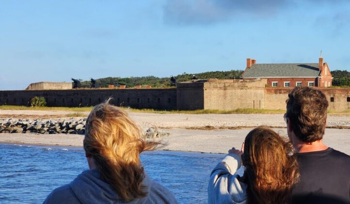 Three people stand near the water, looking toward a historic brick fort with cannons and a nearby building under a blue sky. The sandy shore and stone wall are visible in the background.
