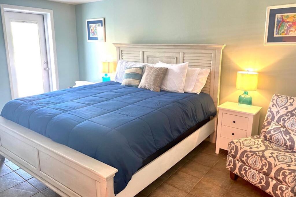 A cozy bedroom with a white bed frame, navy blue comforter, assorted pillows, two turquoise nightstands with yellow lamps, an upholstered armchair, and framed artwork on light blue walls. Natural light enters from a door.