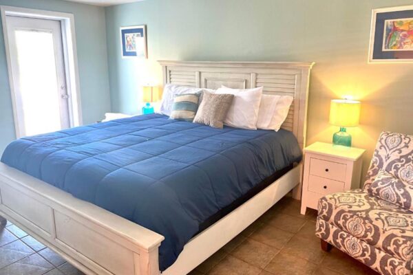 A cozy bedroom with a white bed frame, navy blue comforter, assorted pillows, two turquoise nightstands with yellow lamps, an upholstered armchair, and framed artwork on light blue walls. Natural light enters from a door.