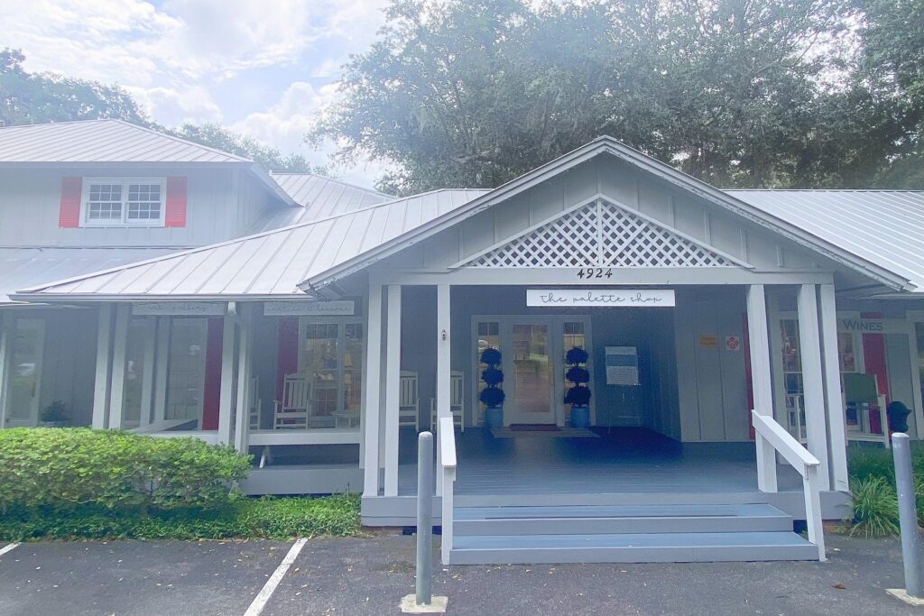A light gray building with a covered porch, white trim, lattice details, and red accents. A sign above the entrance reads the nettles shop with the number 4924. There are rocking chairs and plants on the porch.