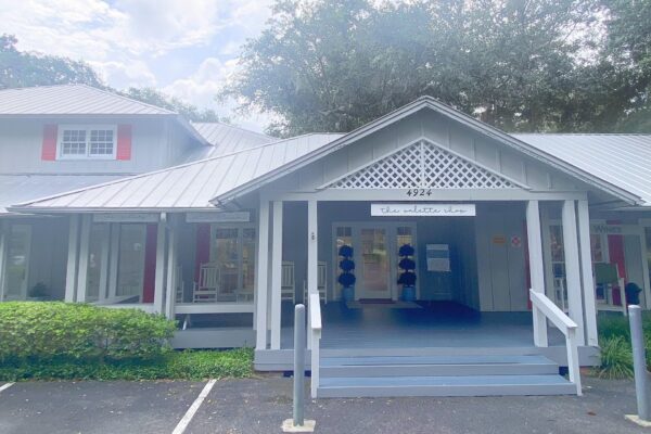 A light gray building with a covered porch, white trim, lattice details, and red accents. A sign above the entrance reads the nettles shop with the number 4924. There are rocking chairs and plants on the porch.