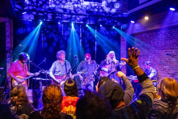 A band performs onstage under blue and purple lights, with guitarists and a singer. Audience members with raised hands watch and take photos in a lively indoor venue with brick walls.
