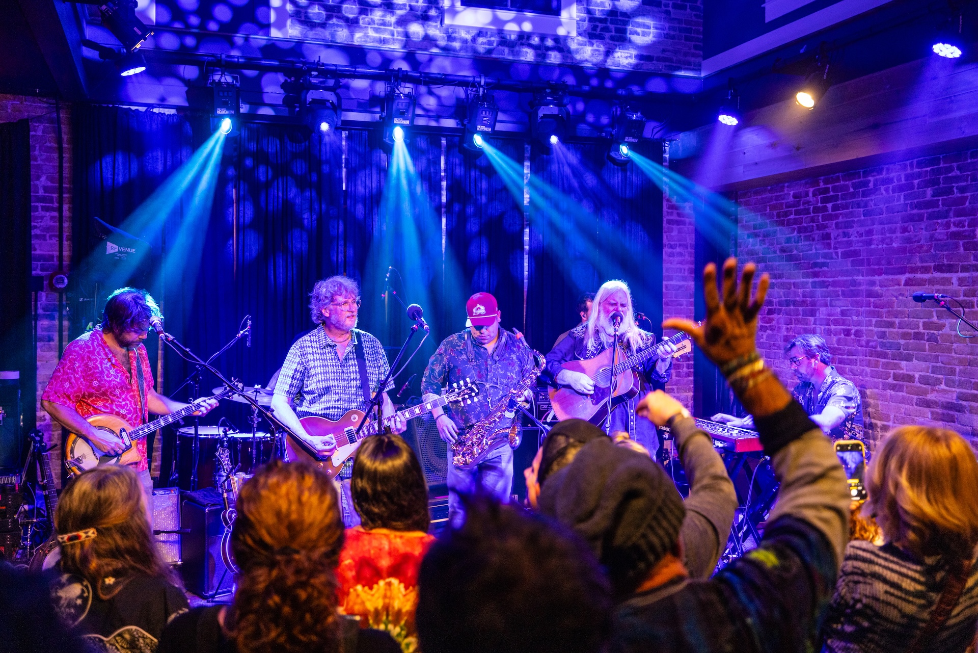 A band performs onstage under blue and purple lights, with guitarists and a singer. Audience members with raised hands watch and take photos in a lively indoor venue with brick walls.