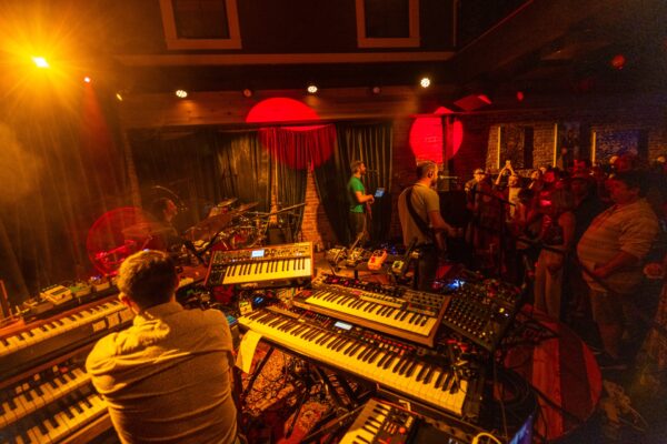 A band performs on stage surrounded by keyboards and music equipment, with warm yellow and red lights. An audience stands close, watching the musicians in an intimate indoor venue with exposed brick walls.