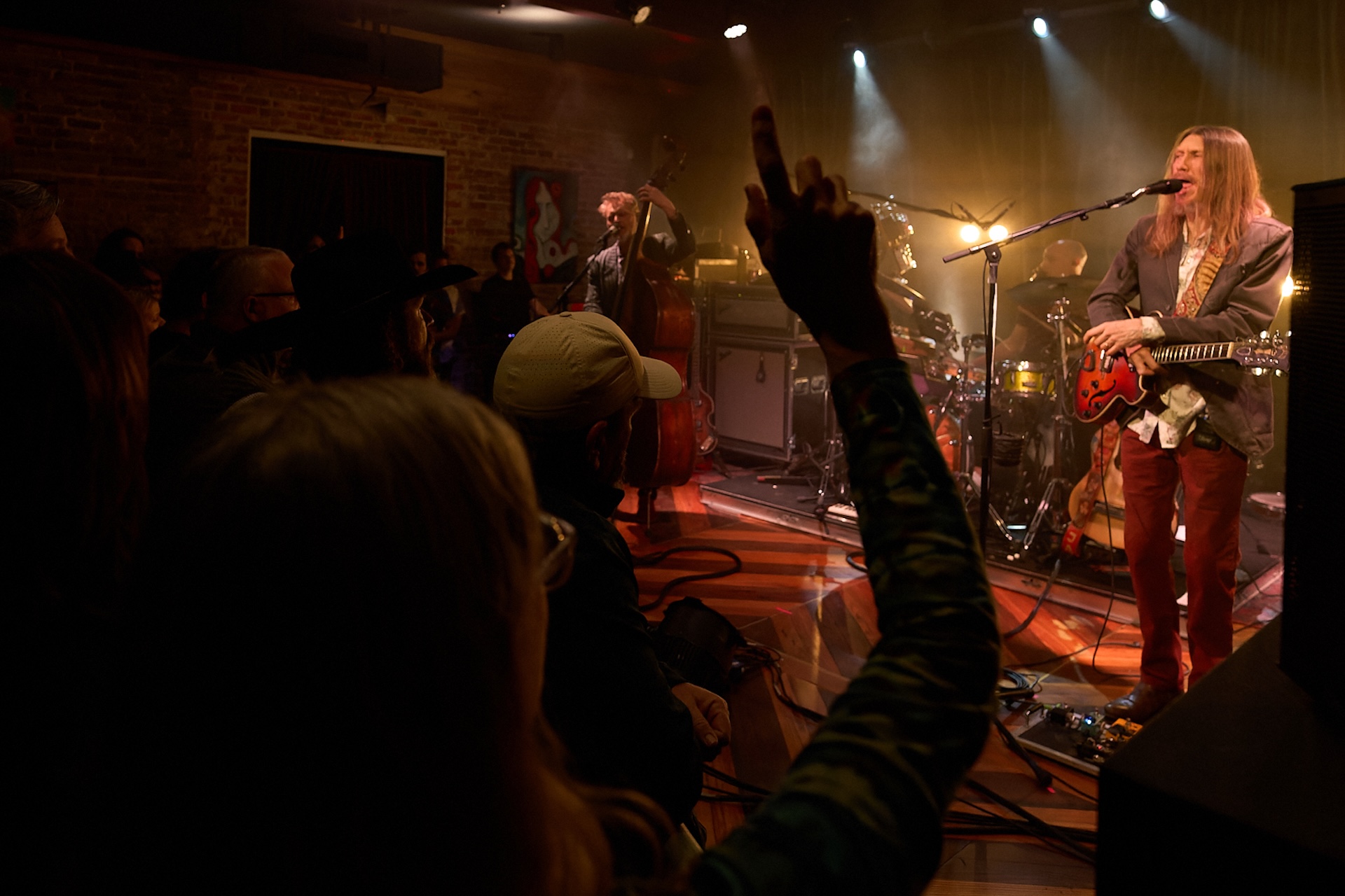 A band performs on a dimly lit stage with a guitarist singing into a microphone. In the foreground, a person in the audience raises a middle finger toward the stage. Other people watch the band, filling the venue.