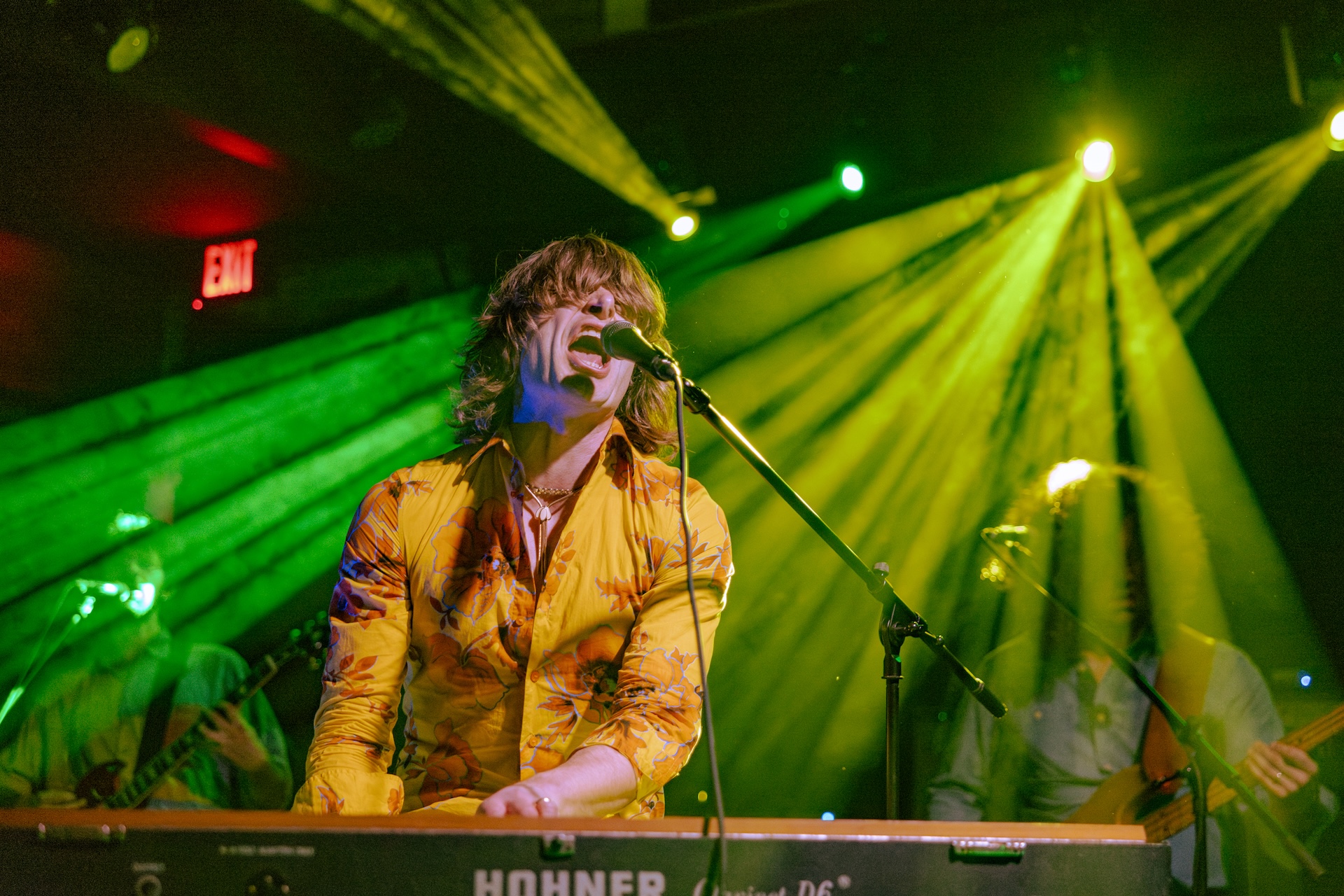 A musician in a yellow floral shirt passionately sings and plays a keyboard on stage, surrounded by dramatic green and yellow stage lights, with band members performing in the background.