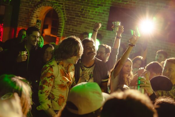 A lively crowd enjoys a party in a dimly lit venue with exposed brick walls. People are smiling, raising drinks, and mingling under green and yellow lights, creating a festive atmosphere.