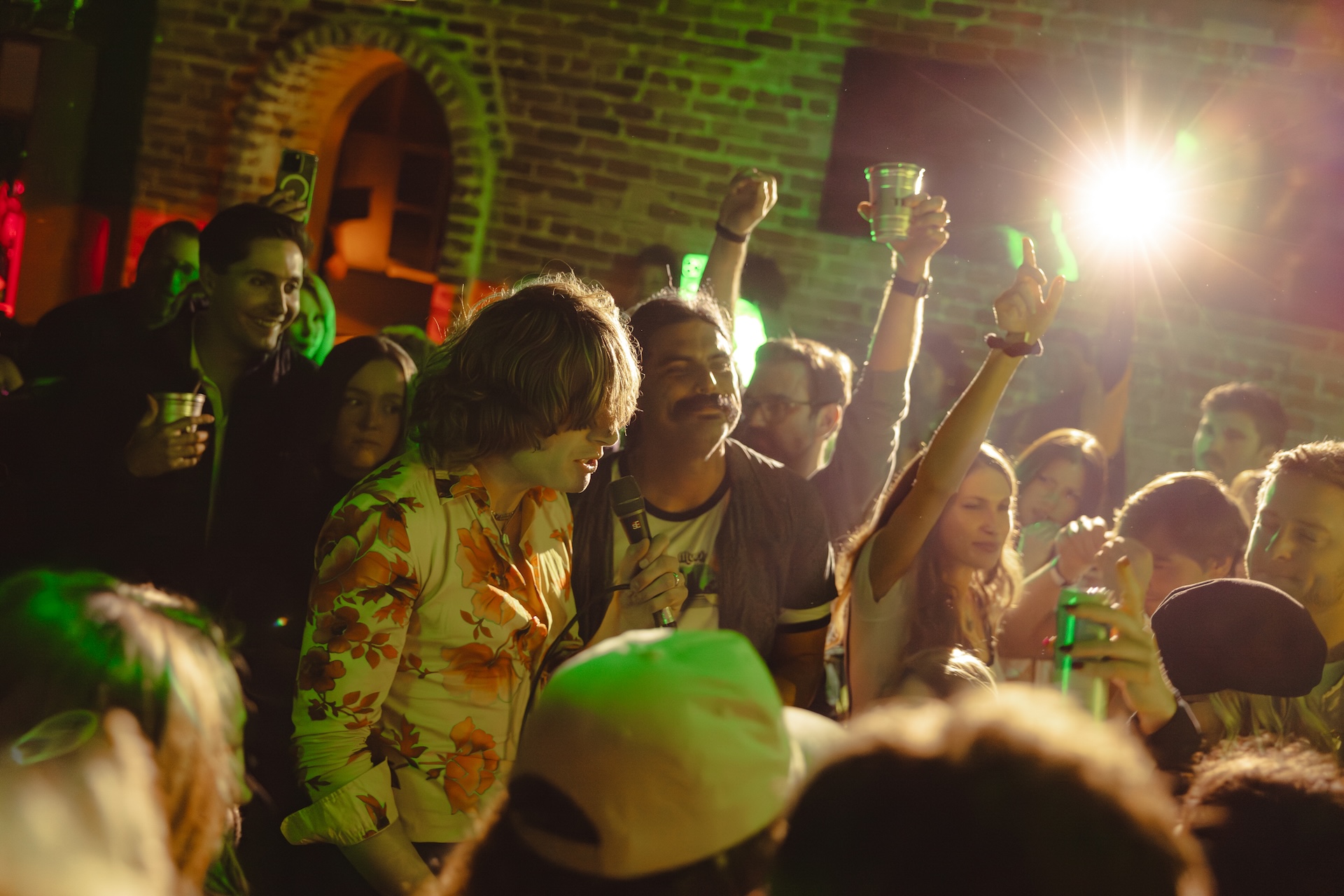 A lively crowd enjoys a party in a dimly lit venue with exposed brick walls. People are smiling, raising drinks, and mingling under green and yellow lights, creating a festive atmosphere.