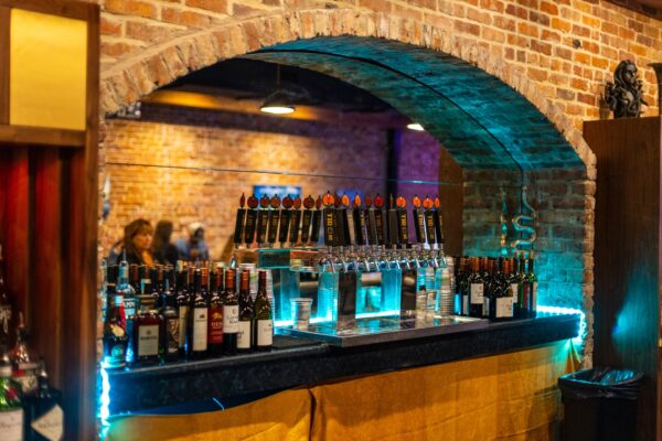 A bar setup under a brick archway with several beer taps, bottles of wine, and plastic cups on the counter. The background features exposed brick walls and people seated at tables.