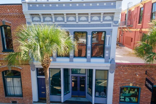 A historic two-story building with light blue and white trim stands between red brick buildings. Large windows and a palm tree partially obscure the facade. Sunlight brightens the scene from above.