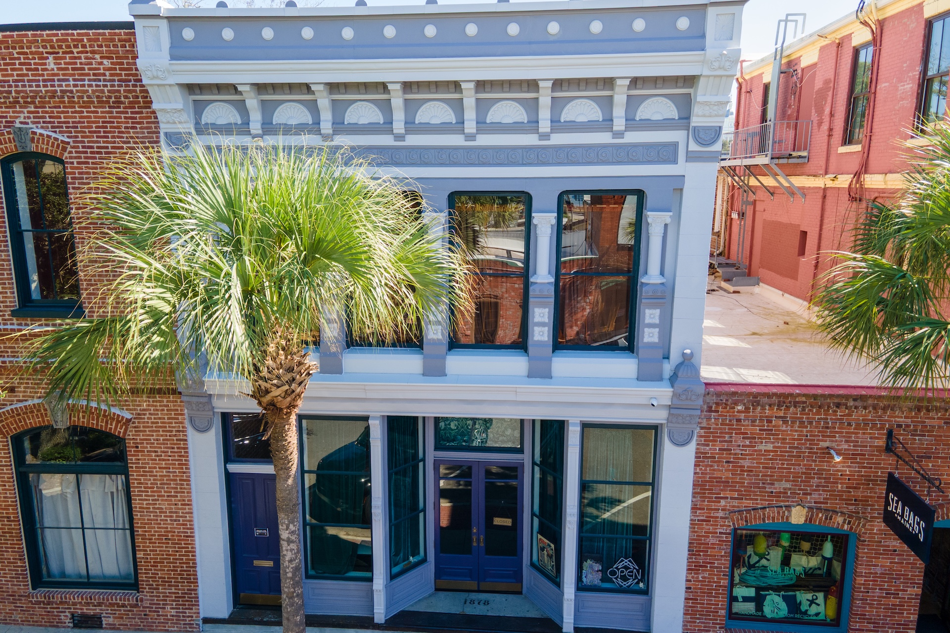 A historic two-story building with light blue and white trim stands between red brick buildings. Large windows and a palm tree partially obscure the facade. Sunlight brightens the scene from above.
