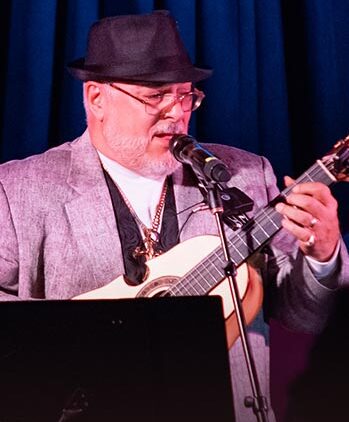 A man wearing a gray suit, black hat, and glasses plays a guitar and sings into a microphone on stage, with dark blue curtains in the background.