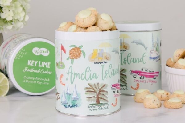 Decorative tins labeled “Amelia Island” and “Key Lime Shortbread Cookies” are filled with cookies. The tins feature colorful beach-themed illustrations. Some cookies are also displayed in a small bowl.