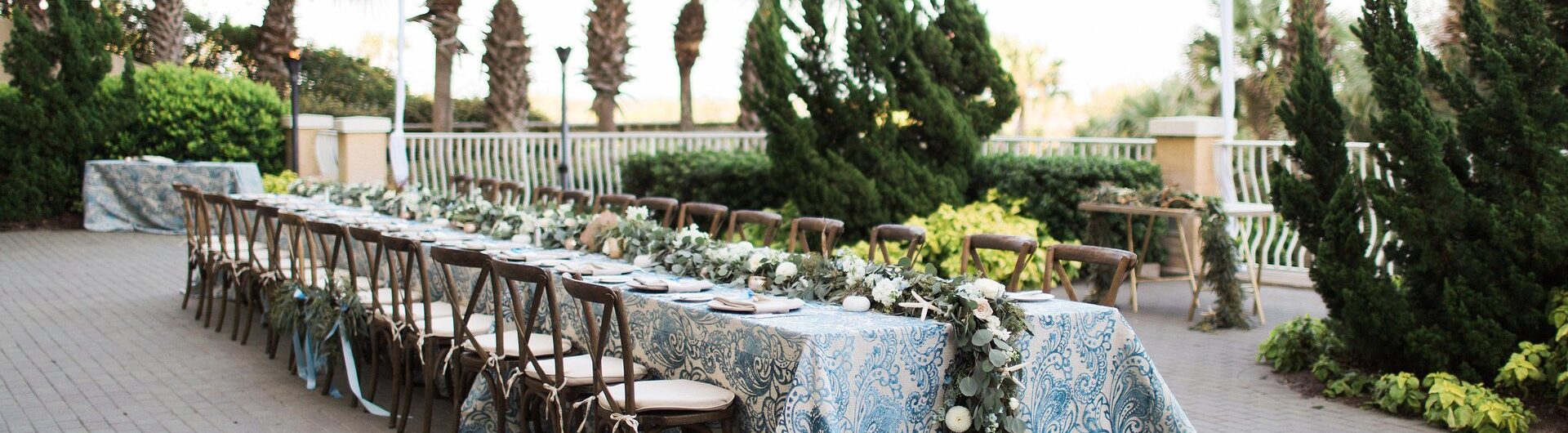 Long outdoor table with blue-patterned tablecloth, floral garland centerpiece, and wooden chairs, set on a patio surrounded by palm trees and string lights.