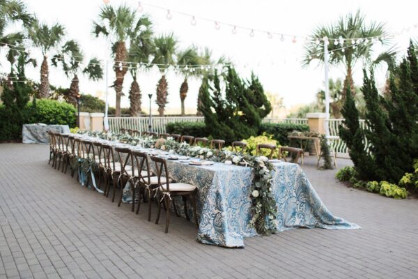 Long outdoor table with blue-patterned tablecloth, floral garland centerpiece, and wooden chairs, set on a patio surrounded by palm trees and string lights.