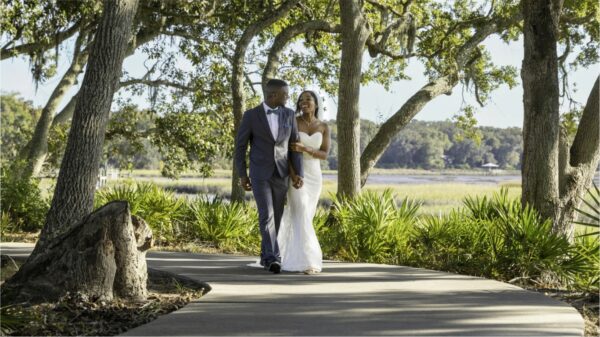 A bride and groom walk hand in hand along a tree-lined path on a sunny day, smiling at each other. Lush greenery and distant water are visible in the background.