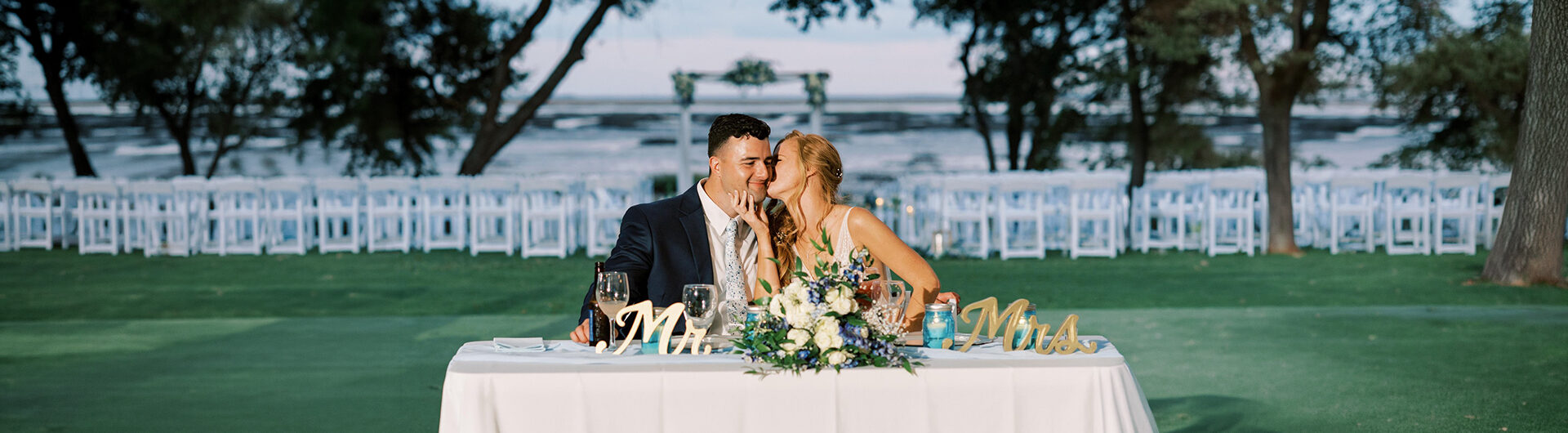A bride and groom sit at a decorated table outdoors, sharing a kiss. Rows of empty white chairs and a scenic waterfront are visible in the background, creating a romantic setting.