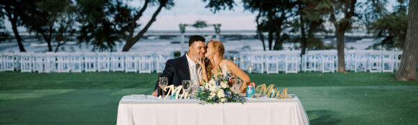 A bride and groom sit at a decorated table outdoors, sharing a kiss. Rows of empty white chairs and a scenic waterfront are visible in the background, creating a romantic setting.