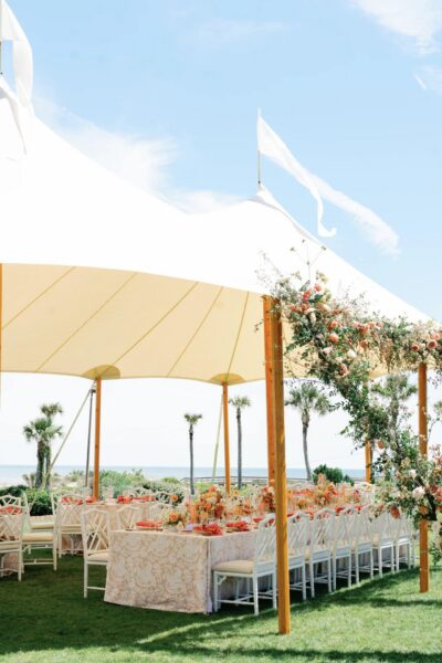 An outdoor event setup under a cream-colored tent, featuring long tables with floral arrangements and white chairs on a grassy lawn, with palm trees and the ocean in the background.
