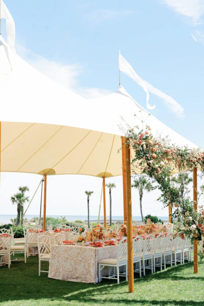An outdoor event setup under a cream-colored tent, featuring long tables with floral arrangements and white chairs on a grassy lawn, with palm trees and the ocean in the background.