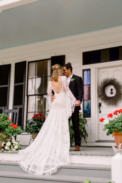 A bride and groom stand closely together on a porch, holding hands and smiling. The bride wears a lacy white dress, and the groom is in a suit. Flowers and greenery decorate the steps and railing.