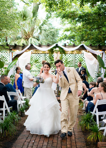 A newlywed couple walks down an outdoor aisle, smiling and celebrating, surrounded by seated guests. The bride wears a white dress and the groom wears a tan suit. Green plants and string lights decorate the venue.