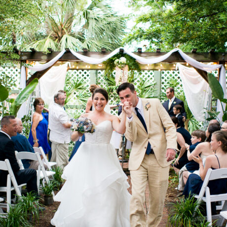A newlywed couple walks down an outdoor aisle, smiling and celebrating, surrounded by seated guests. The bride wears a white dress and the groom wears a tan suit. Green plants and string lights decorate the venue.